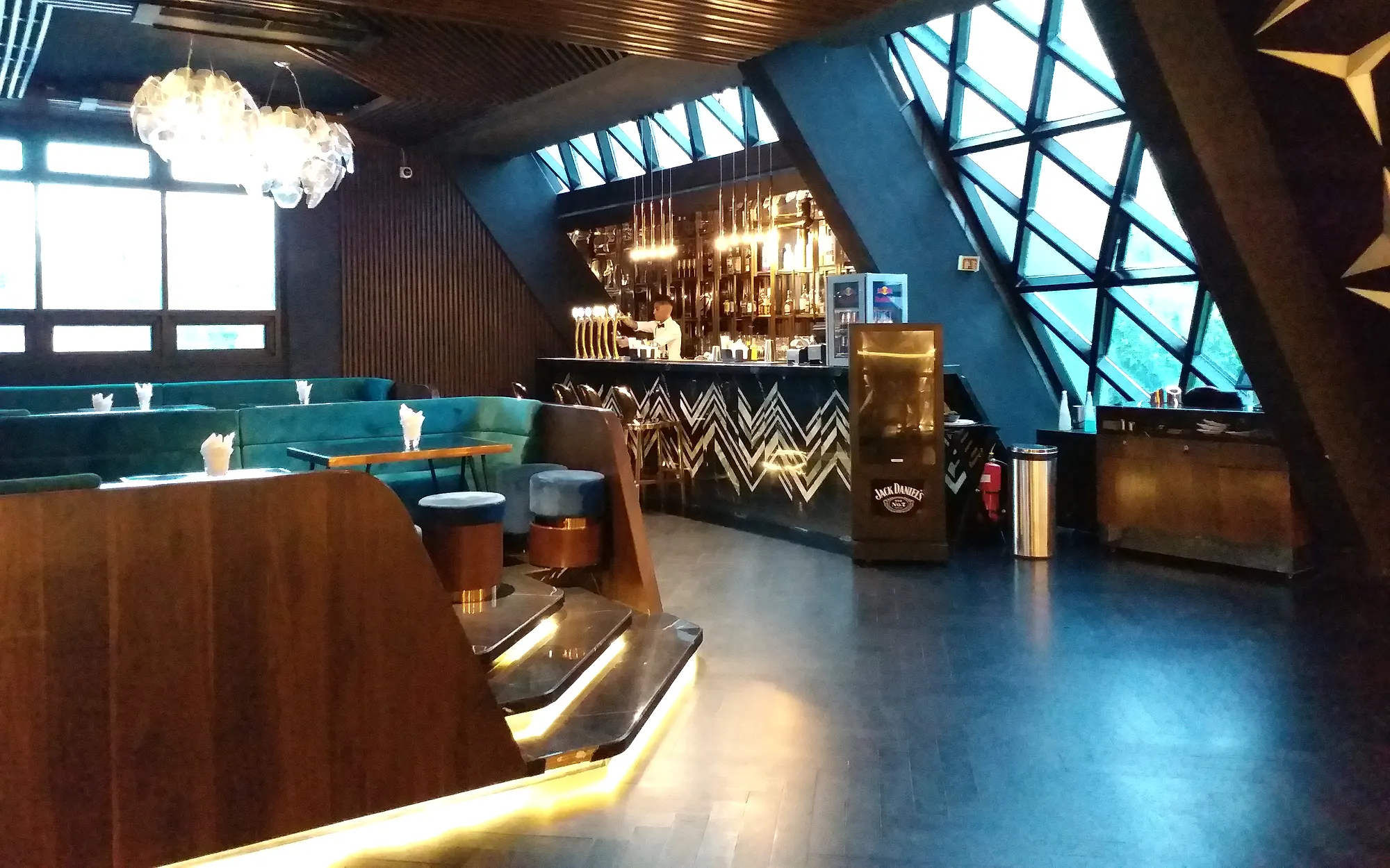 The Pyramid Brewhouse bar at Arena — brass taps and a chevron bar front set under the building's triangulated glass wall, with teal velvet booths in the foreground.
