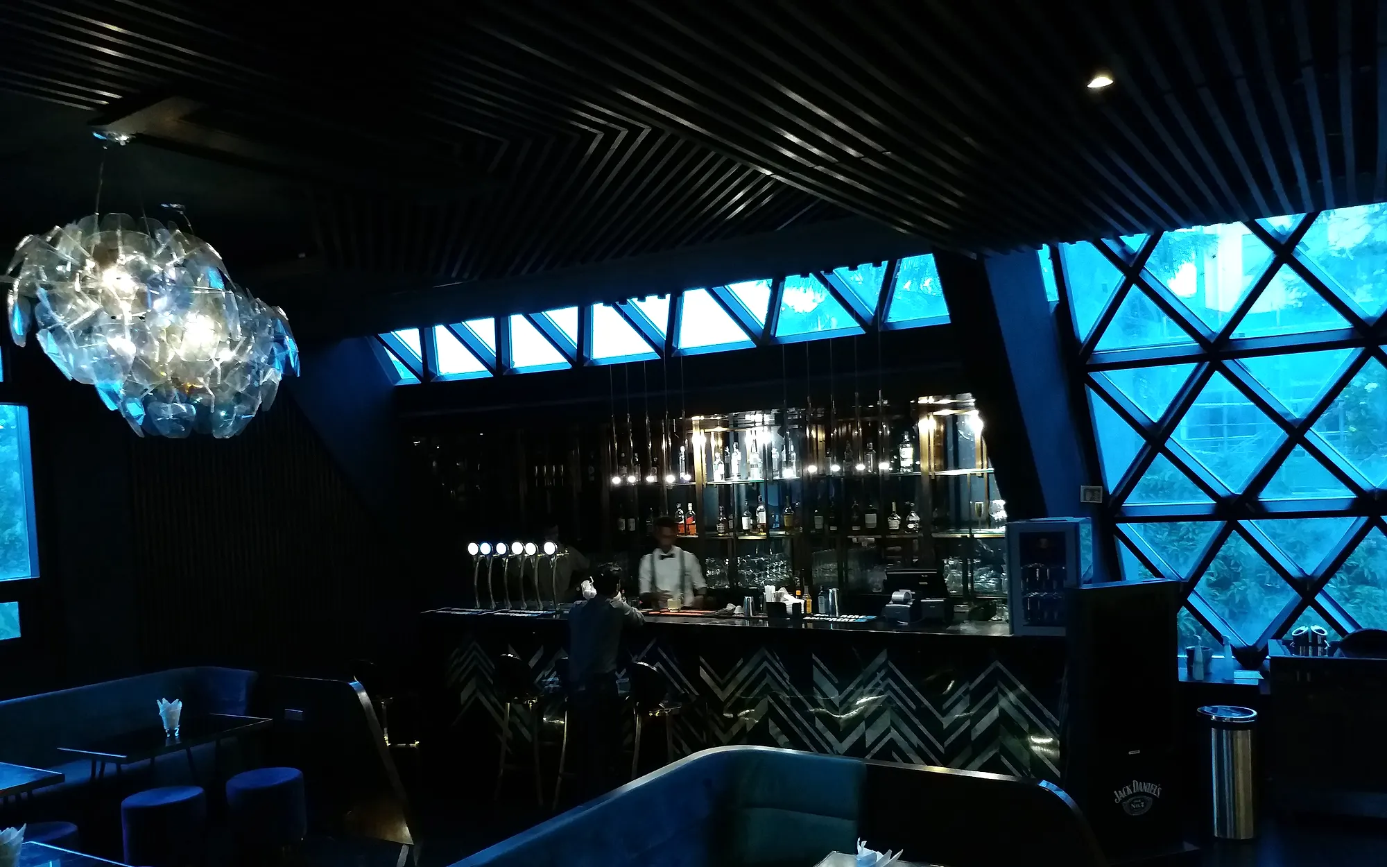 Arena's interior at dusk — daylight coming blue through the diamond-latticed glass of the pyramid, the bar lit warm gold at the far end.