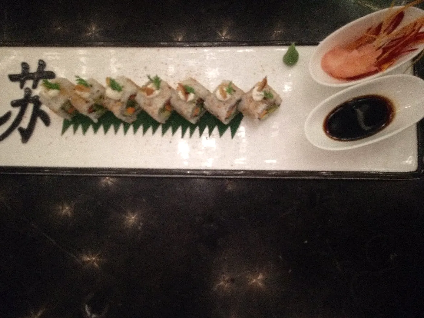 A maki roll plated in a line on a long white slate with wasabi and a dish of soy, alongside a prawn.