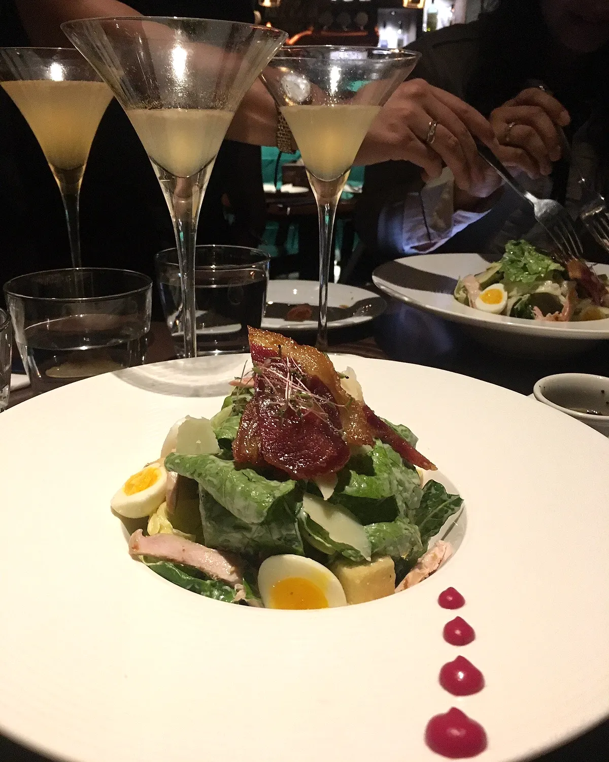A composed salad with quail eggs, shaved cheese and crisped bacon, served on a wide white bowl with martinis behind it.